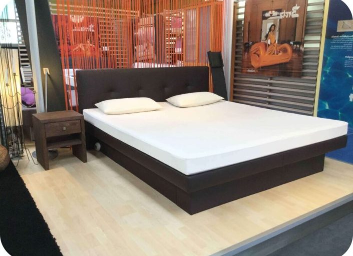 Wasserbetten Hannover - Modernes Schlafzimmer-Display mit einem großen Boxspring-Wasserbett mit weißer Matratze und zwei Kissen auf einem dunklen Bettrahmen. Daneben steht ein Nachttisch aus Holz mit Lampe, ergänzt durch orangefarbene Fadenvorhänge und gerahmte Kunstwerke an der Wand.