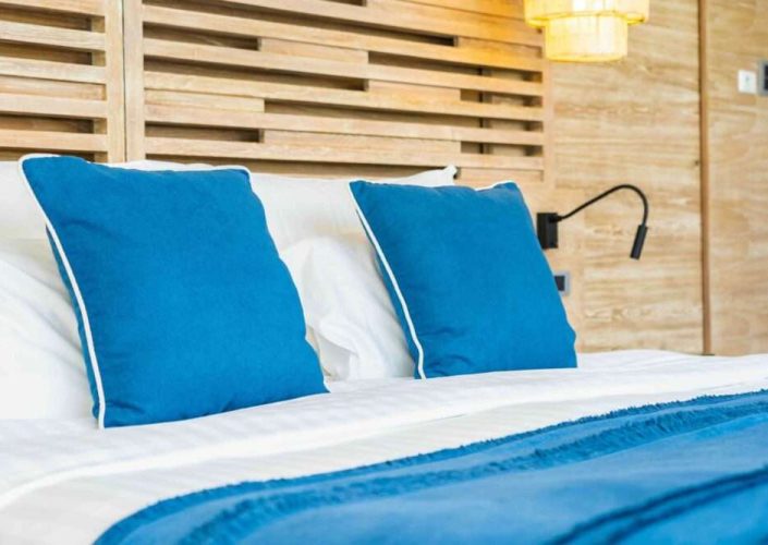 Wasserbetten Hannover - Im Vordergrund steht ein ordentlich gemachtes Bett mit zwei blauen Kissen und einer blauen Decke. Das Bett mit den weißen Laken bietet einen Hauch von Luxus vor einer modernen Holzwandgestaltung. Eine an der Wand montierte Leselampe ziert die Seite. Erleben Sie gehobenen Komfort mit unserem exklusiven Wasserbetten-Service. - Wasserbetten kaufen